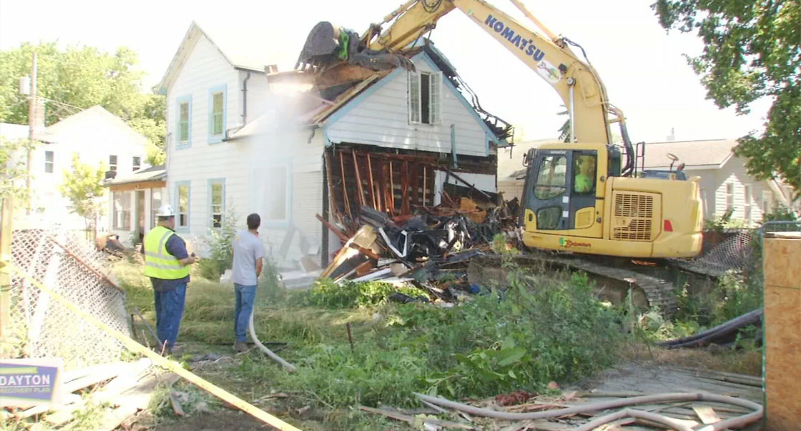 Construction workers using a large Komatsu machine demolish a white house