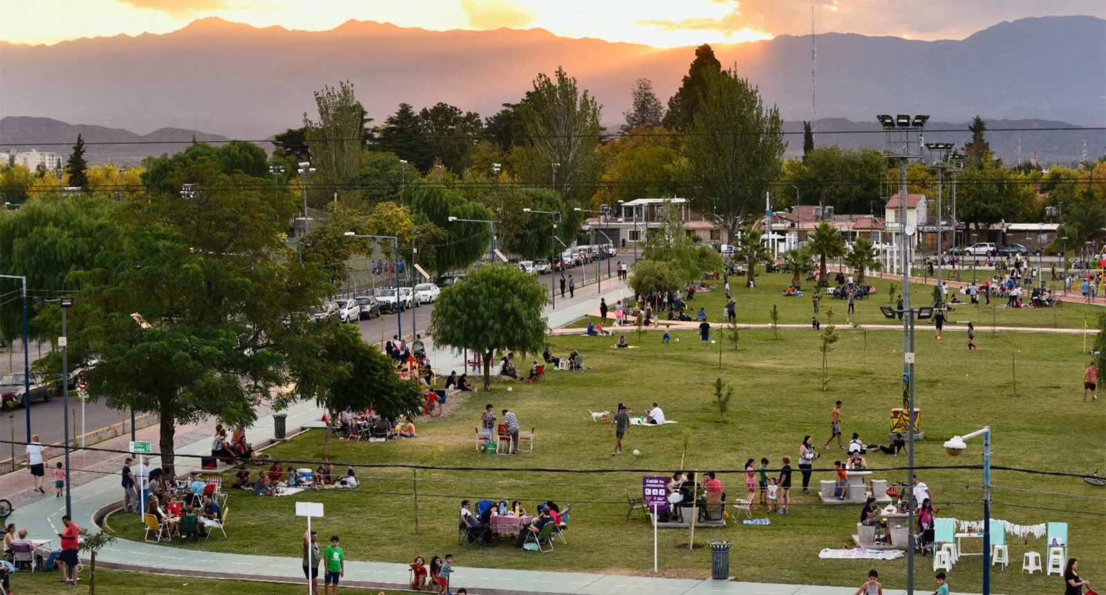 A park in Godoy Cruz is busy with people talking, sitting, and walking with green trees, and beautiful mountains in the background