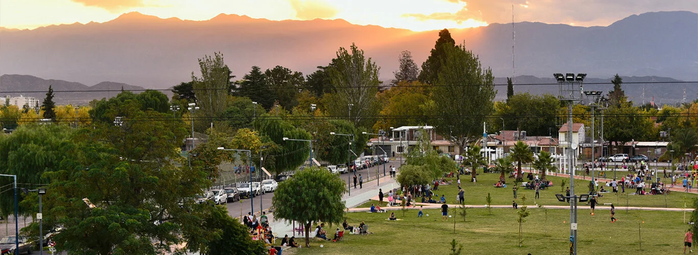 A park in Godoy Cruz is busy with people talking, sitting, and walking with green trees, and beautiful mountains in the background