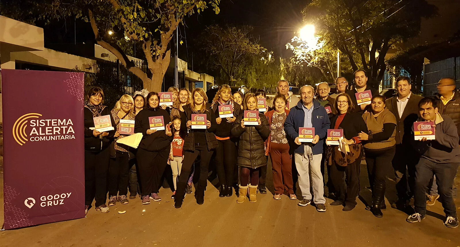 A group of people gather at night outside of a sign reading Sistema Alerta Comunitaria in Godoy Cruz