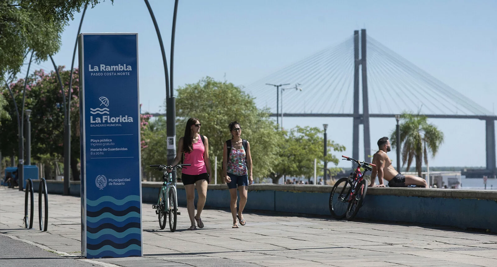 Two women walk on a road near the water with a sign reading La Rambla nearby.