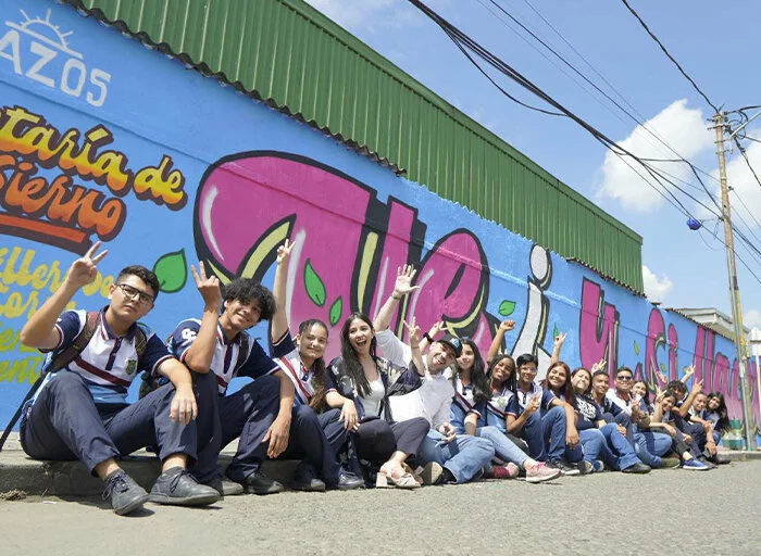 A row of people sitting down next to a mural making the peace sign with their fingers or waving at the camera in Palmira.