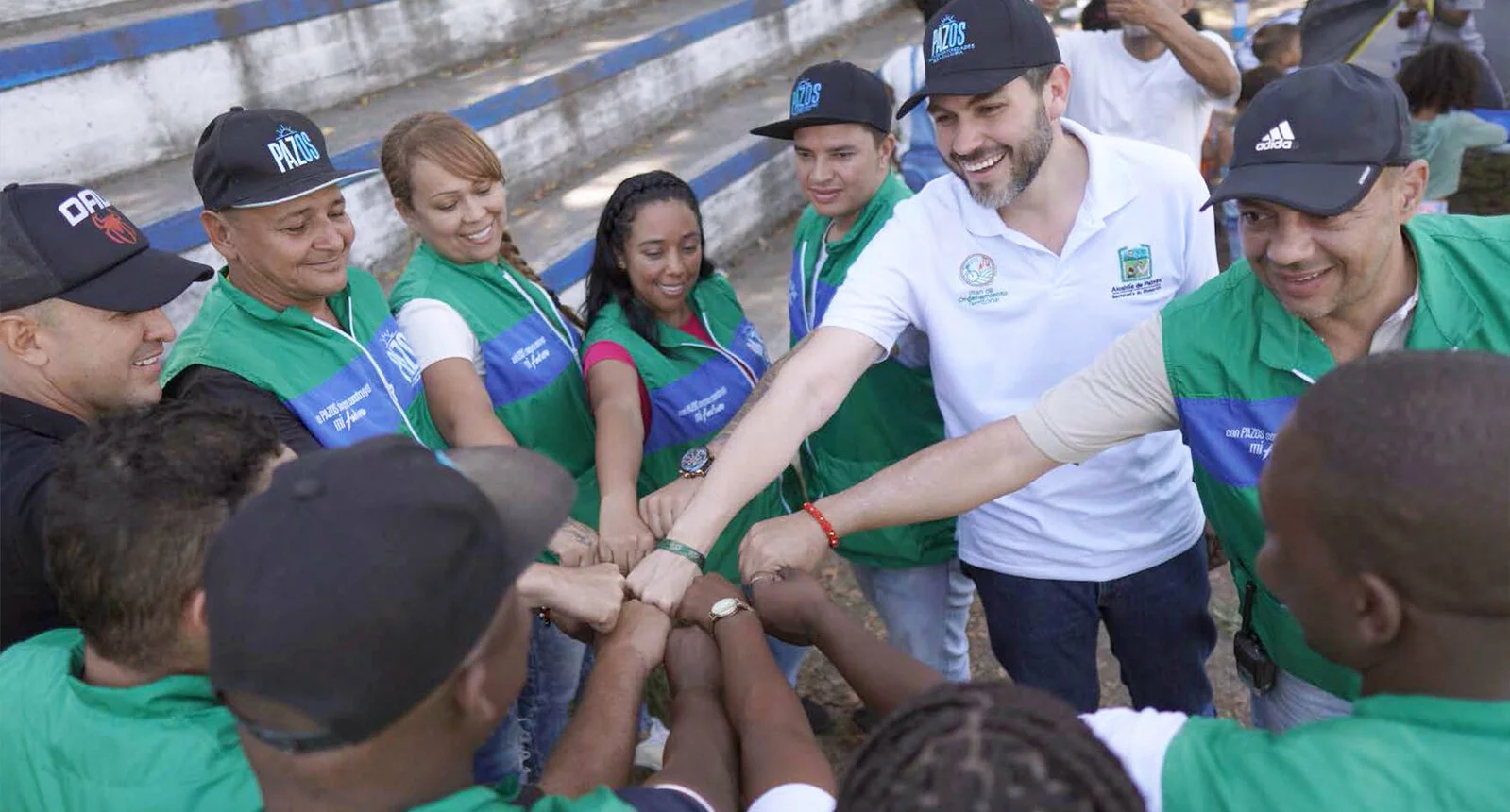 A group of people wearing PAZOS hats and vests stand in a circle holding their fists togethers in a fist bump with smiles