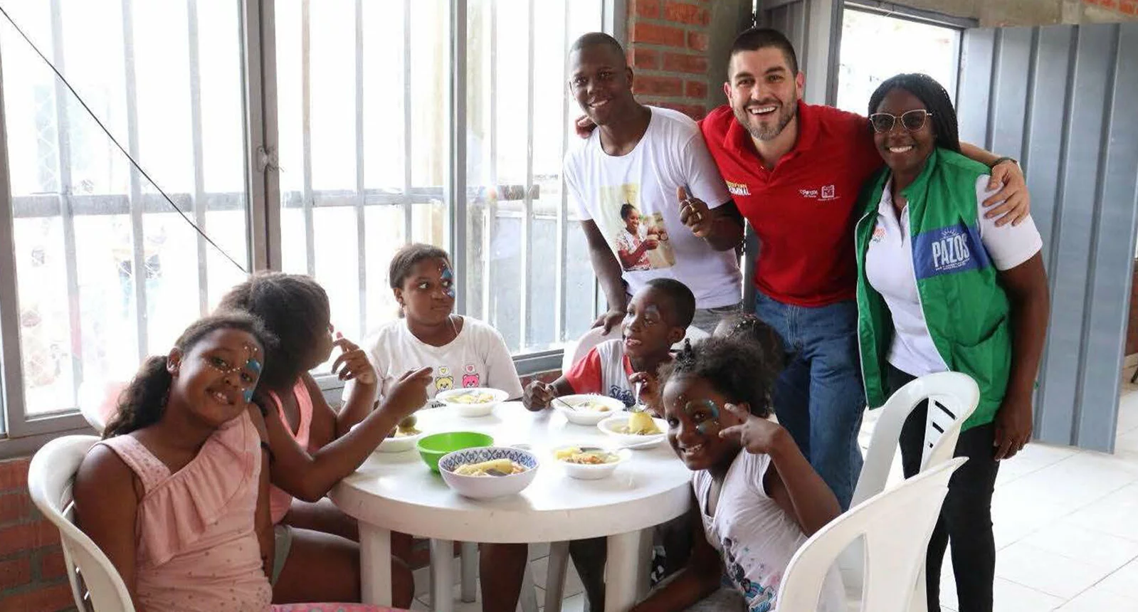 Three people stand smiling (two from PAZOS team) with six children at a table