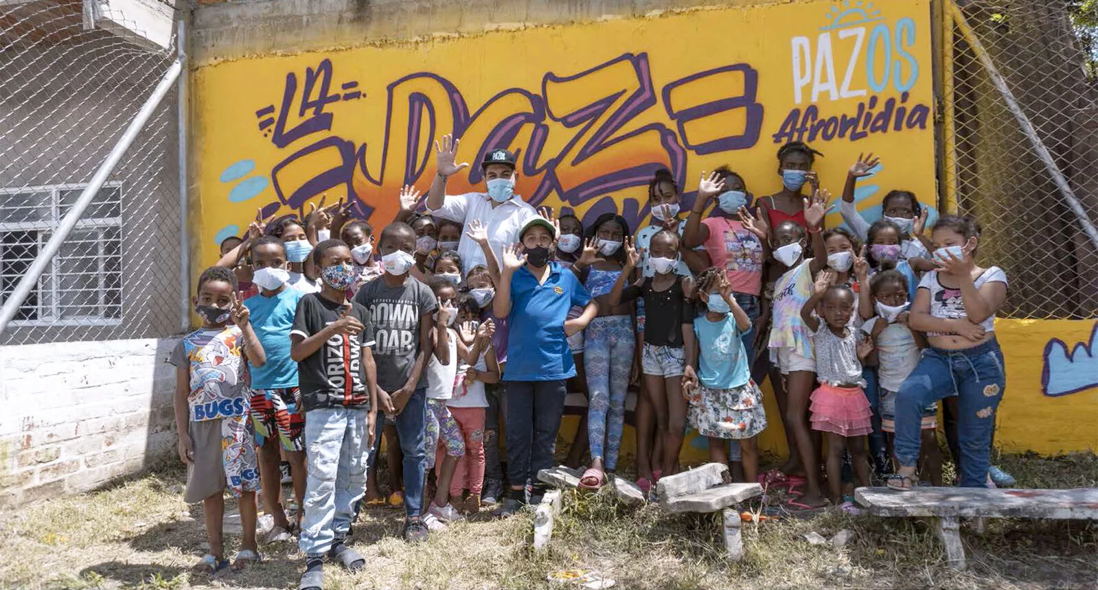 Children wearing masks stand in front of a mural that says PAZ