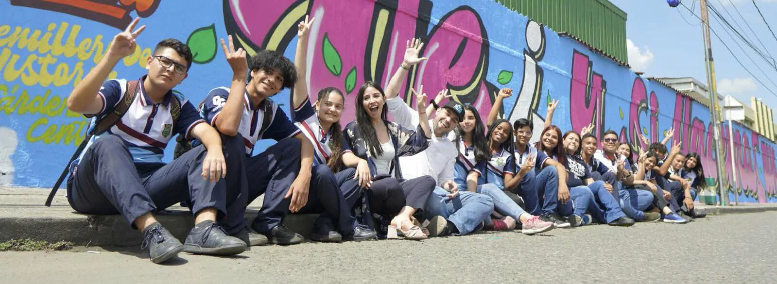 A group of people in Palmira sit on the ground together in front of a mural holding up peace signs with their fingers