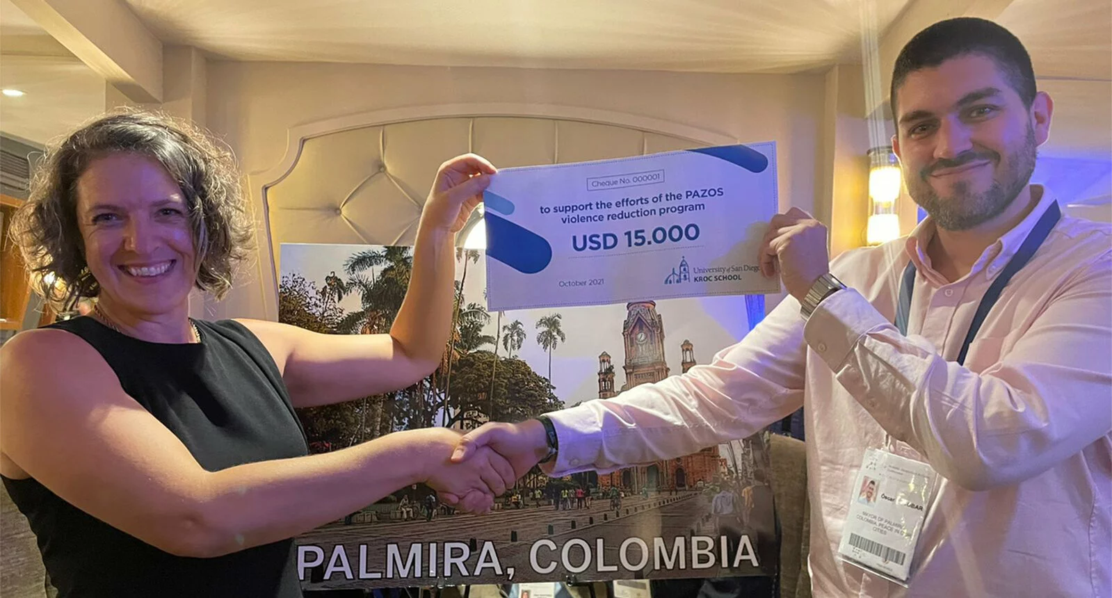 Two individuals hold up a large image of a check and shake hands. Check reads USA 15.000 to support the efforts of the PAZOS violence reduction efforts